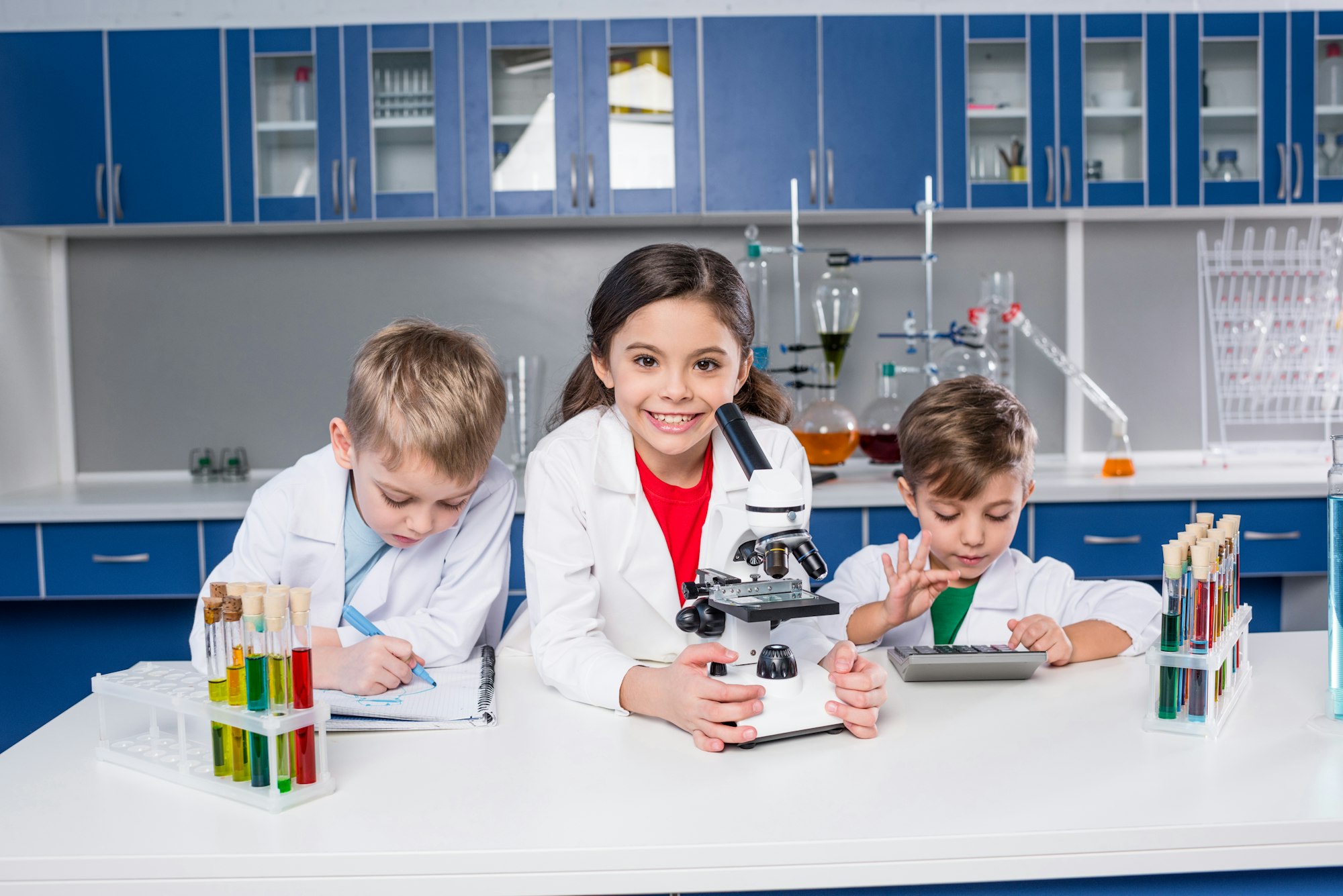 three-kids-in-chemical-laboratory-making-notes-and-using-microscope-and-calculator.jpg